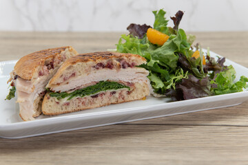 Turkey panini sandwich showing layers of provalone, bacon, mozzarella, and arugula served with a salad on a ciabatta bread bun