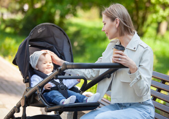 Fototapeta na wymiar Beautiful Caucasian mother sits on a bench with baby in stroller outdoor