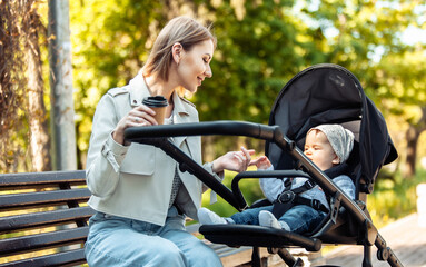 Beautiful Caucasian mother sits on a bench with baby in stroller outdoor