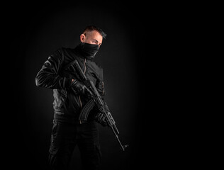 A man in dark clothes holds an automatic carbine in his hands. The face is covered with a mask, hands are in gloves. A figure with a weapon on a dark back