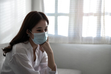 Close-up of a sad Asian patient woman wearing a face mask at home or hospital alone on a sofa...
