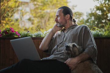 One handsome bearded gentle middle-aged man working on laptop sitting on an open terrace with his dog at home during sunset