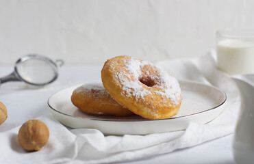A pair of donuts in a plate on a gray table. Next to it is a towel, a nut and a glass of milk.