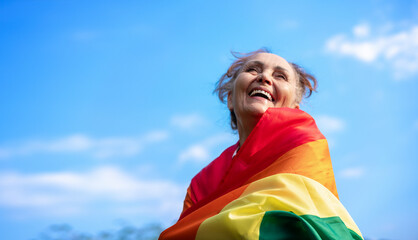 Beautiful mature woman with a charming smile holding a rainbow LGBT flag in her hands, gay and lesbian rights on a blue sky background - obrazy, fototapety, plakaty
