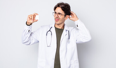 young hispanic man feeling puzzled and confused, scratching head. physician with pills bottle...