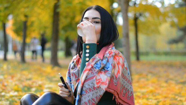 Beautiful stylish young girl portrait brunette drinks hot coffee and smokes an electronic cigarette in the autumn city park