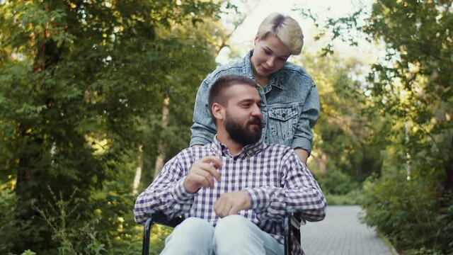 Disabled man in wheelchair with his girlfriend on walk park, they talk and smile, front view. Caucasian couple walks park, woman carries disabled man in wheelchair