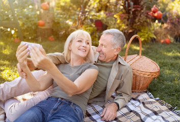 Fototapeta premium Modern elderly spouses taking selfie on smartphone, having picnic and resting in garden at sunny autumn day