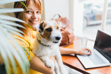 Portrait of young girl student sititng at table with computer and dog at work place. Pretty amazing female freelancer working at cafe with jack russell terrier.