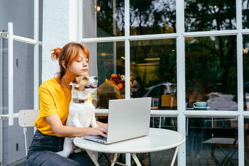 Lovely pet concept. Girl work with laptop, plays with a dog. Redhead woman hugging with dog at cozy cafe outdoor. Shelter a mongrel at home. Life style. College People Study Learning Reading Notes. - obrazy, fototapety, plakaty
