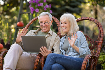 Modern grandparents. Happy senior spouses having video call via laptop, relaxing in wicker chairs...