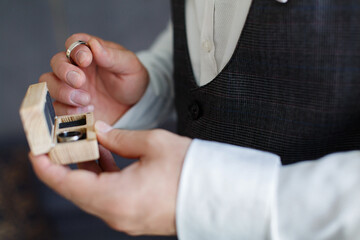 Men's hand with two gold wedding rings in the wooden box close up.
the groom in black suit holds the wedding rings of white gold on palm on background. wedding ceremony. wedding day. bride and groom