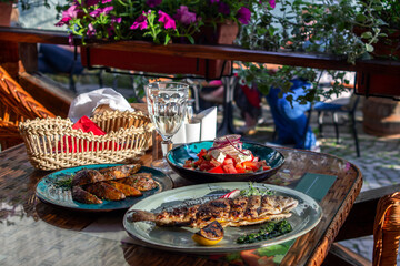 Fototapeta premium server table with a freshly prepared three-course lunch on the summer terrace of the cafe. Grilled trout, grilled potatoes, fresh tomato, pepper and onion salad, a glass of white wine. Tasting.