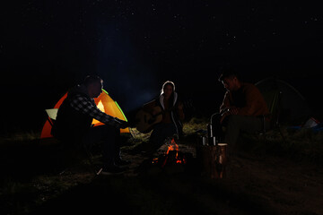 Naklejka premium Group of friends with guitar near bonfire and camping tent outdoors at night
