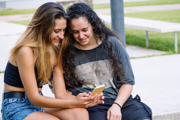 Two girlfriends sitting outdoors looking at the smartphone.