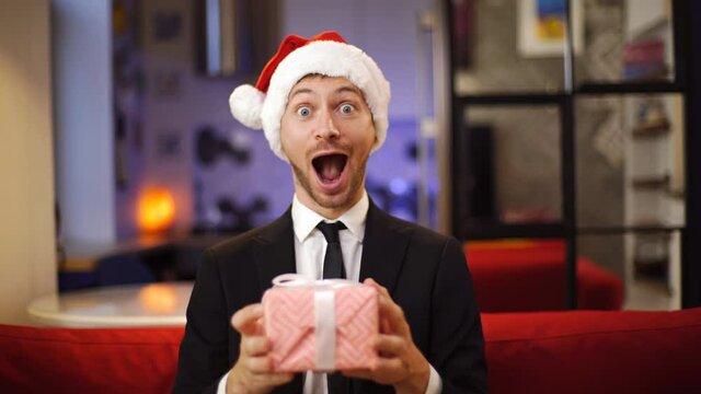 Surprised businessman dressed suit in red Santa hat holding a Christmas gift box