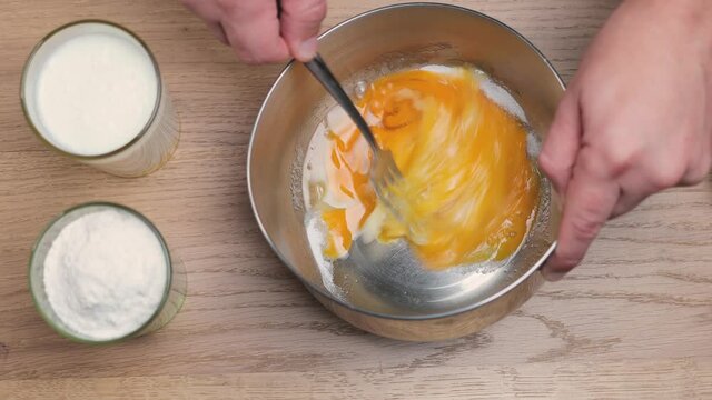 Male hand with fork blends the broken chicken eggs with a sugar in a metal bowl, cooking concept, top view