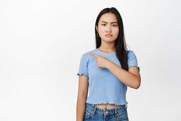 Angry asian woman pointing finger left, pouting and frowning upset, standing in blue tshirt over white background