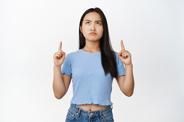 Moody asian girl pointing and looking up, frowning with angry face expression, standing in blue tshirt over white background
