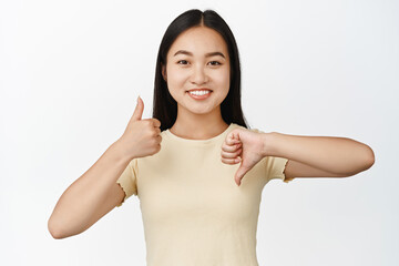 Portrait of smiling asian girl shows thumbs up and thumbs down, like and dislike gesture, standing...
