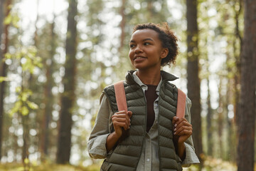 Waist up portrait of young African-American woman with backpack enjoying hiking in forest lit by...