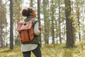 Back view portrait of young African-American woman with backpack enjoying hiking in forest lit by...