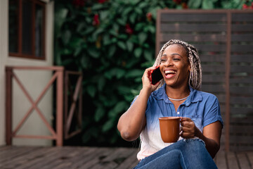 Mujer madura afroamericana con cabello con trenzas sonriendo mientras habla por celular y toma una taza de café o té