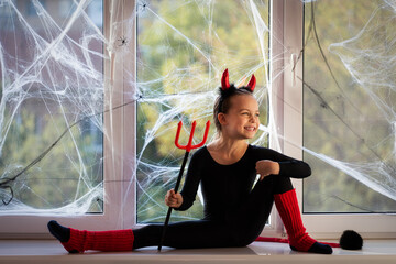 Little girl gymnast in costume of little devil smiling and playing about on window sill