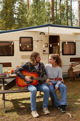Vertical full length portrait of happy young couple playing guitar while enjoying camping outdoors...
