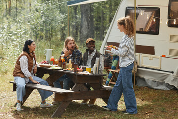 Full length view at diverse group of friends enjoying picnic outdoors at campsite with trailer van