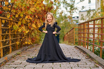 Fototapeta premium Happy laughing little girl on autumn trail in long magic black fashion dress. Halloween in gorgeous princess dress. Retro smile. Place for text. Laughing funny child girl in witch costume. road bridge