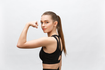 Fototapeta na wymiar Waist up shot of sporty woman raises hand to show her muscles, feels confident in victory, looks stong and independent, smiles positively, stands against grey background. Sport concept