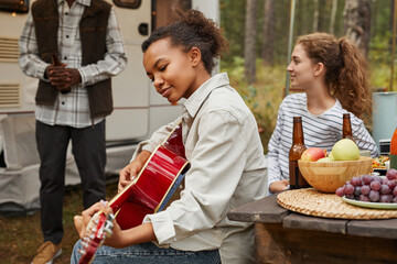 Portrait of young African-American woman playing guitar while enjoying camping outdoors with friends