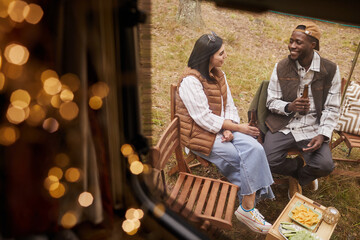 High angle view at young couple relaxing while camping outdoors by van lit by fairy lights, copy space