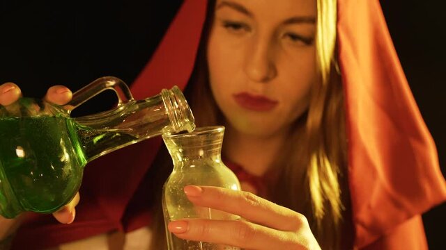 Woman in red cloak pours a green liquid into glass bottle. Witch in red hood prepares potion for Halloween