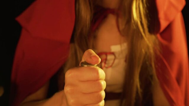 Witch in red hood is pounding ingredients for potion in an iron mortar. Magic rite at night