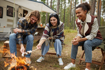 Group of young people roasting marshmallows while enjoying camping with friends in forest, copy...