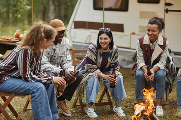 Full length portrait of modern young people enjoying camping outdoors and sitting by fire drinking beer