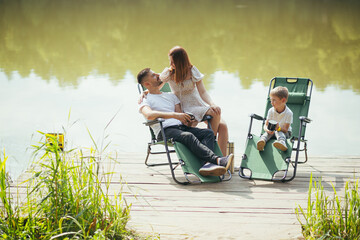 Happy young family with toddler children resting sitting on folding camping chairs over lake on a wooden pier outdoors. spend leisure time together in camp pontoon with kids in nature. vacation forest