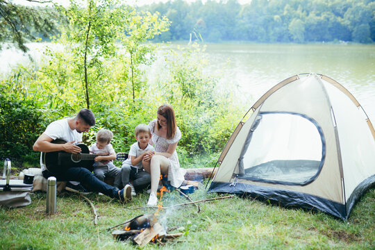 Camping By Lake In Woods. Happy Family Dad Mom And Little Kids Sitting By Fire And Tent In Nature. Spend Leisure Time Together On Vacation. Outdoors. Parents With Children. Father Playing Guitar. Camp