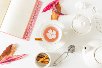 Tea time composition with a cup of tea with a white flower inside, pink and brown leaves, tea pot, open book, cinnamon on white background. Flat lay, top view. Copy space.