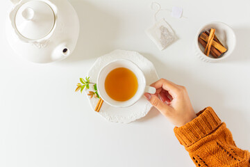 Composition with female hands holding a cup of tea, teapot, cinnamon and tea bag on white background. Flat lay, top view. Slow morning concept.