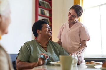 Portrait of smiling young woman assisting female patient during dinner at nursing home, copy space