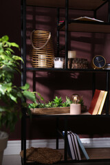 Stylish wicker candle holder, night lamp with incandescent bulb and decor on shelving unit near brown wall