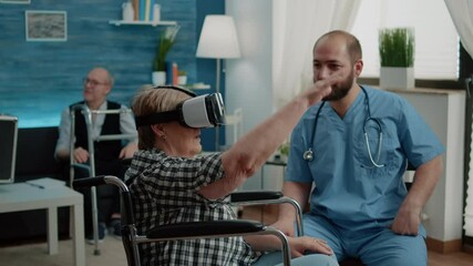 Disabled old woman in wheelchair using vr glasses in nursing home while man nurse explaining virtual reality for healthcare and recovery treatment. Patient with handicap and technology - Powered by Adobe