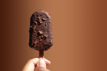 Delicious chocolate and almond coated ice cream skewers on a wooden stick and hold by left hand isolated on a gradient brown background with clipping path and copy space for input text.