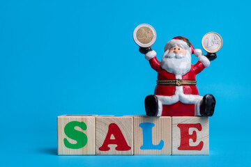 Ceramic toy Santa Claus sits on the word SALE, assembled from colorful letters on children's cubes and holds a 2 euro coin in one hand and 1 euro coin in the other