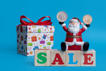 Ceramic toy Santa Claus sits on the word SALE, assembled from colorful letters on children's cubes and holds a 50 American cent coin in one hand and 1 American dollar coin in the other