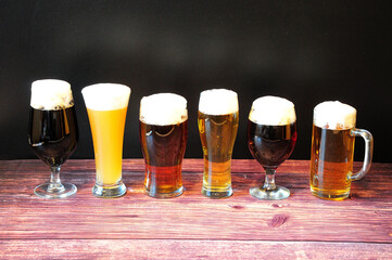 Six different glasses of beer with six different types of beer with froth stand in a row on a wooden table.