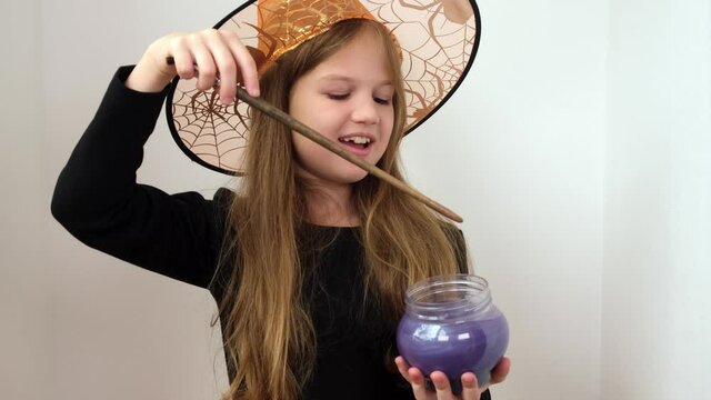 Little girl in orange witch hat is making magic potion. Kid is in costume for Halloween party. Child is stirring a soft purple substance in a cup and terribly laughs.